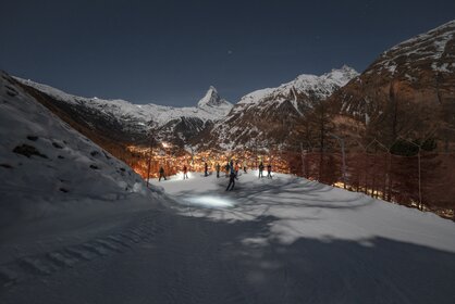Piste in der Dunkelkeit mit Skifahrern, die auf das beleuchtete Zermatt hinunter blicken, im Hintergrund das Matterhorn | © Gabriel_Perren