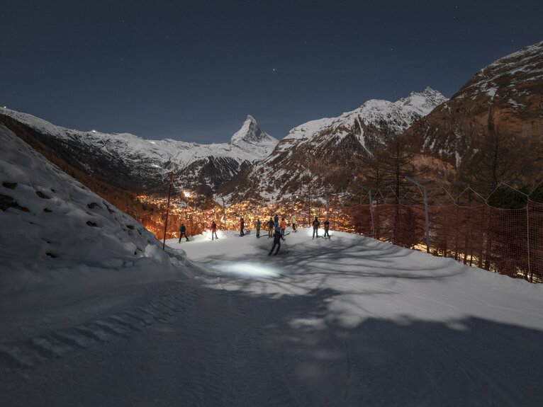 Piste in der Dunkelkeit mit Skifahrern, die auf das beleuchtete Zermatt hinunter blicken, im Hintergrund das Matterhorn | © Gabriel_Perren