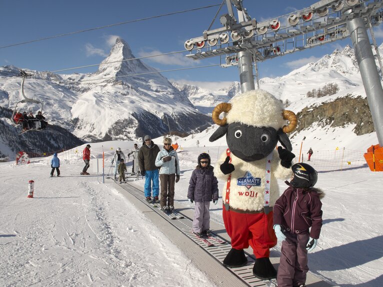 Eine Familie und Wolli stehen auf dem Zauberteppich im Anfängerpark Sunnegga. Im Hintergrund ist die Bergwelt zu sehen.  | © Zermatt Bergbahnen