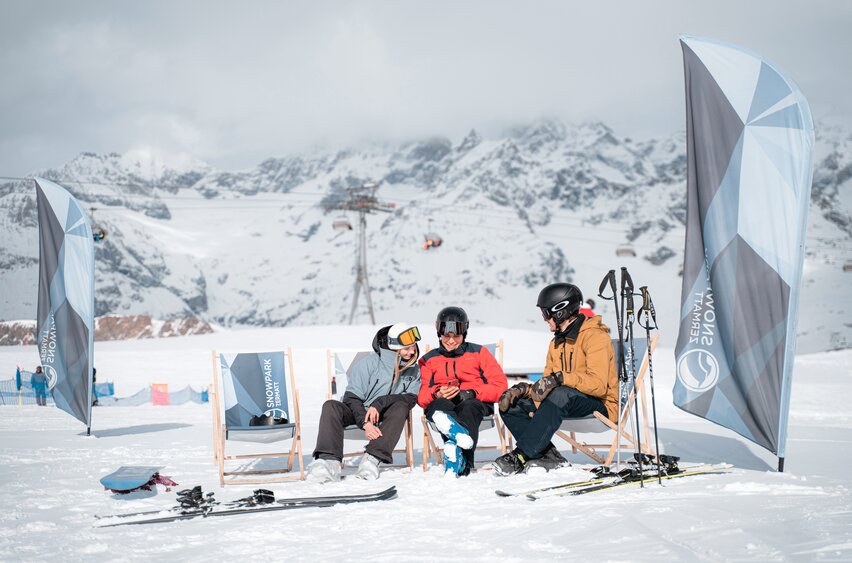 Drei Personen unterhalten sich im Snowpark auf den Liegestühlen.  | © Basic Home Production
