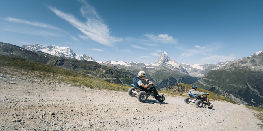 Due uomini sfrecciano lungo il sentiero su mountaincarts con il Cervino sullo sfondo. | © Gabriel Perren