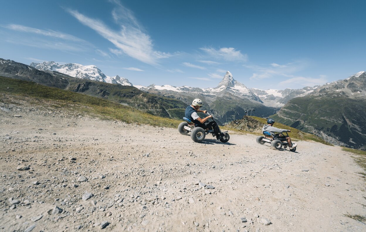 Due uomini sfrecciano lungo il sentiero su mountaincarts con il Cervino sullo sfondo. | © Gabriel Perren