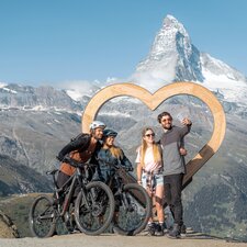 Zwei Wanderer und zwei Biker machen ein Selfie vor dem Holzherz auf Blauherd.  | © Basic Home Production