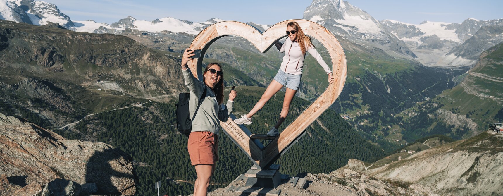 Zwei Frauen machen ein Selfie vor dem Holzherz auf Blauherd. | © Gabriel Perren