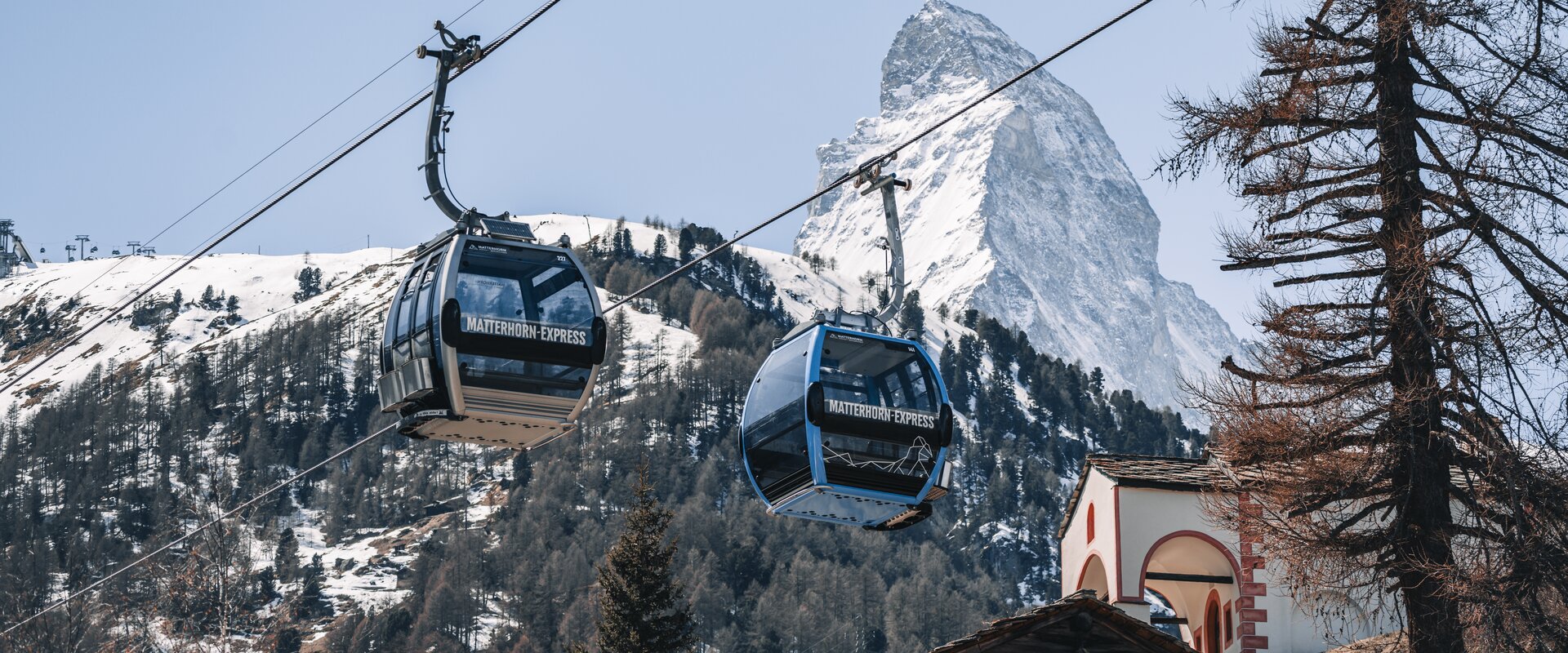 2 Gondeln schweben im Frühling über der Kapelle in Blatten und vor dem weissen Matterhorn.  | © Gabriel Perren 